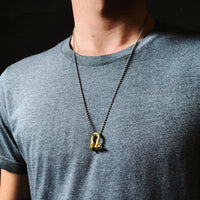 The necklace portion is featured on a model wearing a gray shirt. The brass shackle with discs on the post is strung on a stainless steel ball chain. The bottom of the necklace rests on top of the model's sternum.