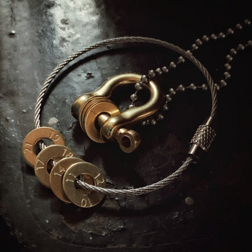 A brass shackle with brass numbered discs strung on the post is strung on a stainless steel ball chain. The shackle is framed by a stainless steel cable with more brass numbered discs strung on it. The numbers are stamped on the discs, evenly spaced around the disc with the bottom of the number oriented towards the hole in the middle of the disc.
